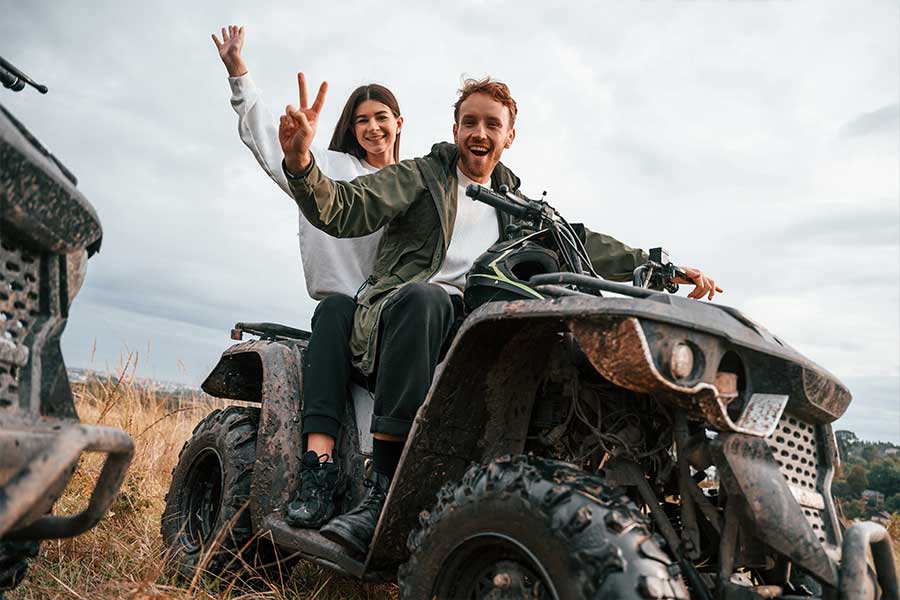 A happy couple riding a muddy ATV, with the woman in the back making a peace sign and the man in front smiling, against an overcast sky.