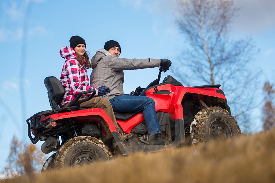 A man and a woman in winter hats and jackets riding a red ATV uphill on a dirt path with bare trees in the background under a blue sky.