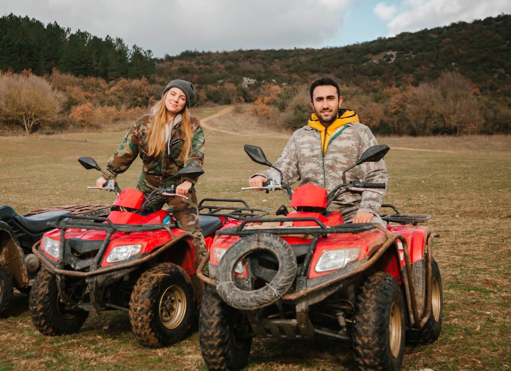A smiling man and woman on red ATVs in a grassy, mountainous landscape with trees.