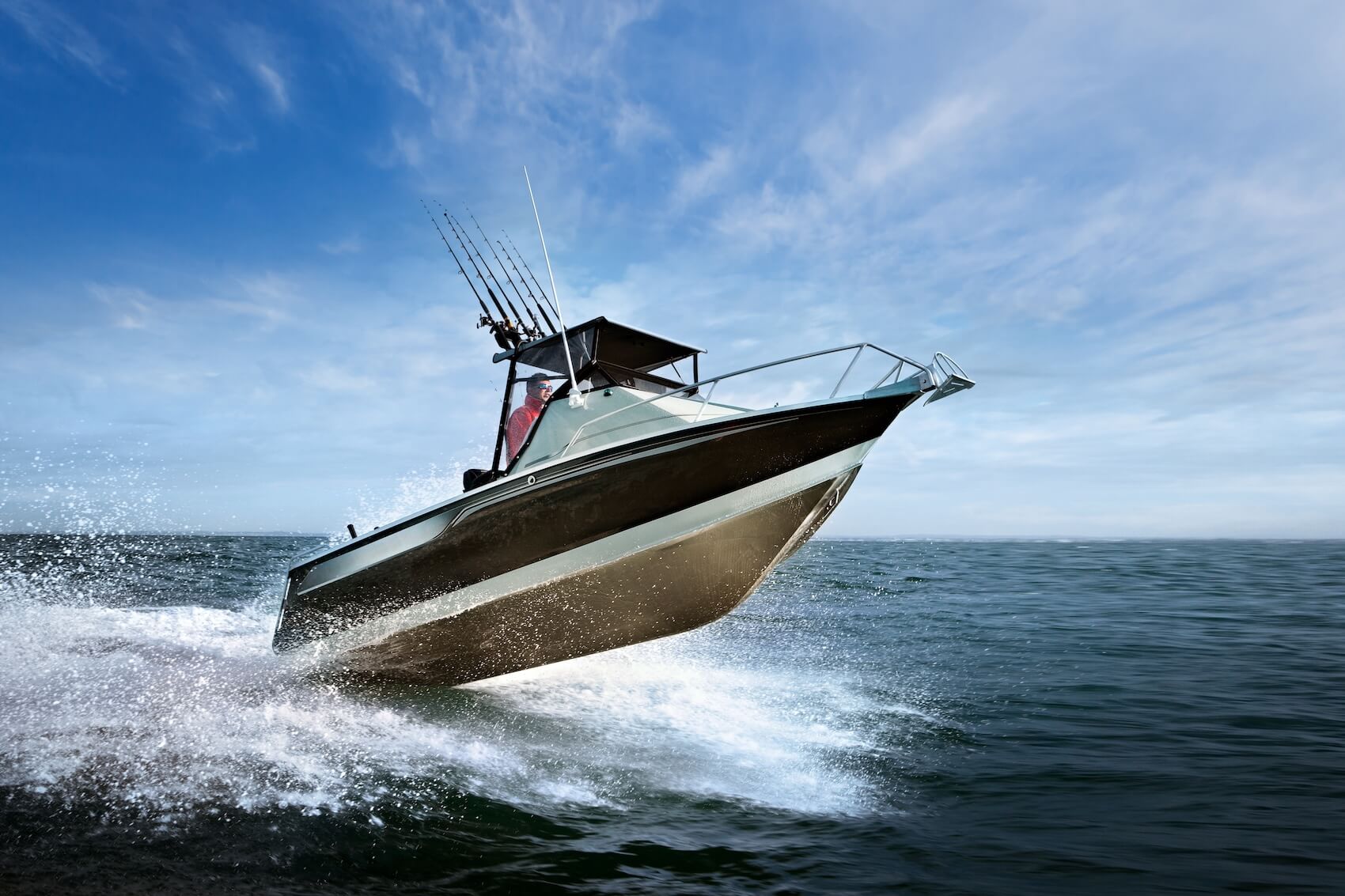 Offshore fishing boat speeding across the ocean, kicking up white spray, under a partly cloudy blue sky.